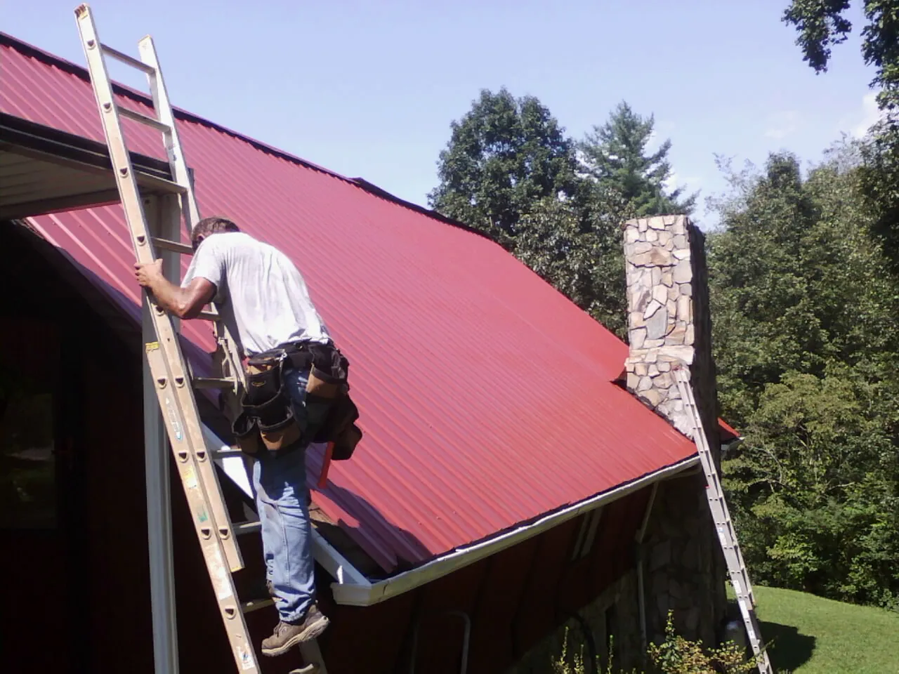 Roofer on a ladder beside a red metal roof and stone chimney — Soffit & Fascia Repair by Roofing Brothers in Crows Landing